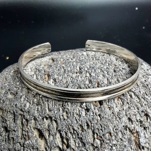 Könnte beinhalten: Ein silbernes Armreif-Armband mit einem doppellienigen Design. Das Armband liegt auf einer strukturierten grauen Oberfläche.