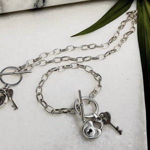 May include: A silver chain necklace and bracelet set with a padlock and key charm. The necklace and bracelet have a toggle clasp.