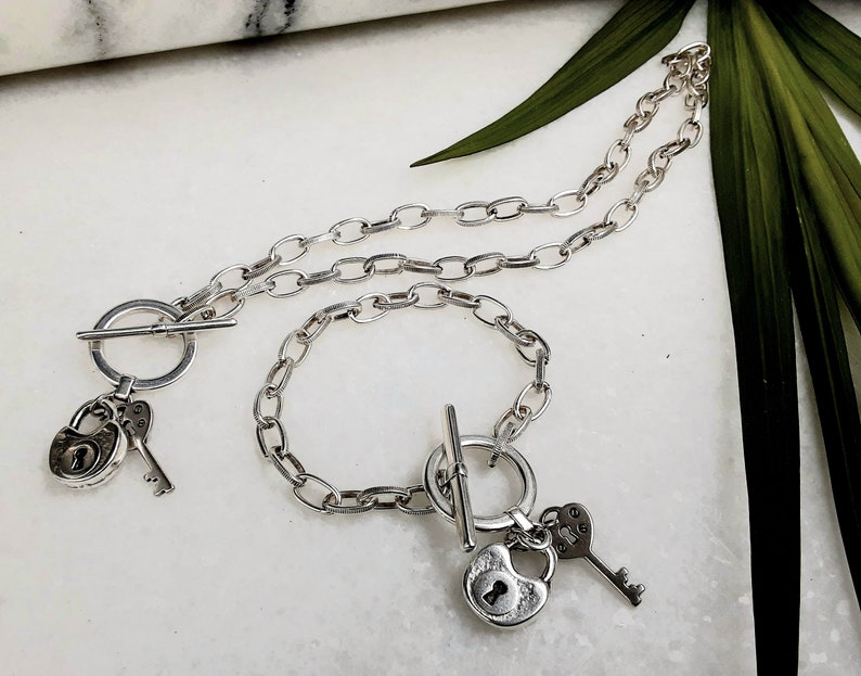 May include: A silver chain necklace and bracelet set with a padlock and key charm. The necklace and bracelet have a toggle clasp.