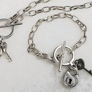 May include: A silver necklace and bracelet set with a padlock and key charm. The necklace and bracelet have a toggle clasp.