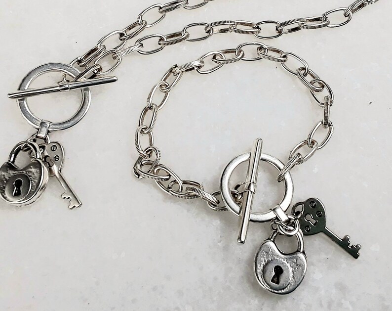 May include: A silver necklace and bracelet set with a padlock and key charm. The necklace and bracelet have a toggle clasp.