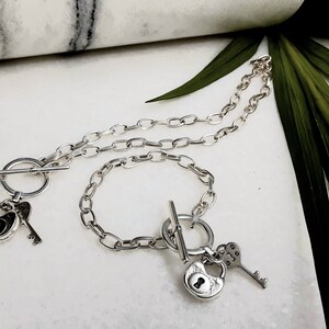 May include: A silver necklace and bracelet set with lock and key charms. The necklace and bracelet feature a toggle clasp.
