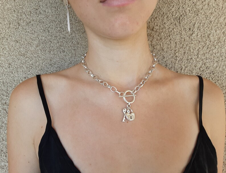 May include: A silver chain necklace with a toggle clasp and a heart-shaped padlock charm with a key attached.