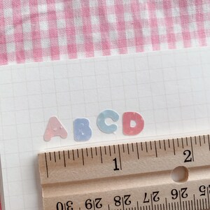 以下が含まれることがあります： パステルカラーの文字ステッカー、A、B、C、Dのクローズアップ。方眼紙に貼られています。木製の定規は、ステッカーの高さが約1.3cmであることを示しています。背景はピンクと白のチェック柄です。