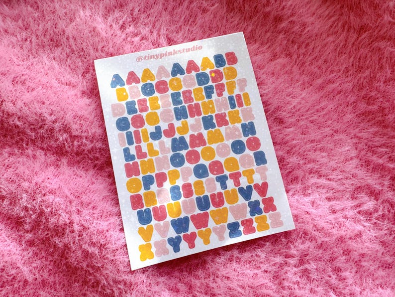 以下が含まれることがあります： ピンク、黄色、青、オレンジなど、さまざまな色の文字が並んだアルファベットステッカーのシート。 ステッカーはグリッドパターンで配置され、白い背景にあります。 上部には「@tinypinkstudio」というテキストがあります。