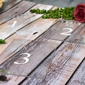 May include: Three clear acrylic table numbers with white text. The numbers are 1, 2, and 3. The table numbers are on a wooden surface.
