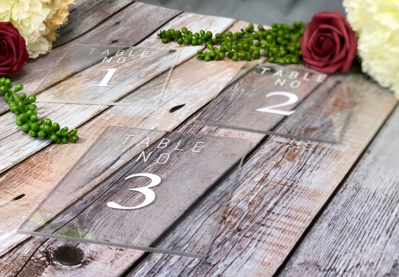 May include: Three clear acrylic table numbers with white text. The numbers are 1, 2, and 3. The table numbers are on a wooden surface.