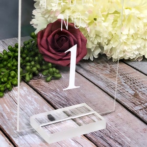 May include: Clear acrylic table number sign with the text "Table No. 1" in white lettering. The sign is sitting on a wooden surface with flowers in the background.