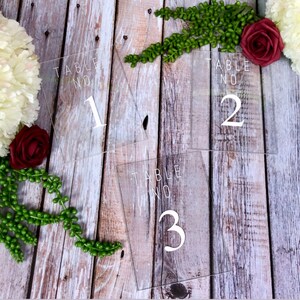 May include: Three clear acrylic table number signs with white text. The signs read "Table No. 1", "Table No. 2", and "Table No. 3".