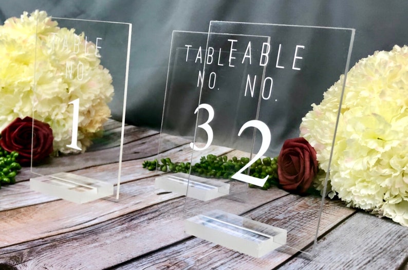 May include: Clear acrylic table number holders with the text "Table No." and the numbers "1", "3", and "2" printed in white. The holders are on a wooden surface with flowers.