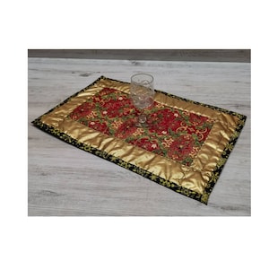 Könnte beinhalten: Ein roter und goldener gesteppter Platzdeckchen mit schwarzem Rand und goldener Einfassung. Ein klares Glas steht auf dem Platzdeckchen.
