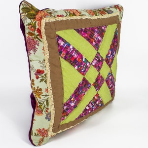 Op de afbeelding: Een vierkant kussen met een patchwork-ontwerp. Het kussen is gemaakt van verschillende stoffen in tinten groen, bruin, roze en paars. Het kussen heeft een bloemenprint aan de achterkant.