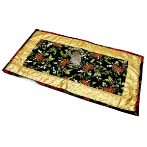 Könnte beinhalten: Ein schwarzer und grüner Tischläufer mit goldener Umrandung und einem roten und grünen Stechpalmenmuster. Der Läufer ist gesteppt und hat eine satinartige Oberfläche. Eine klare Glasvase steht auf dem Läufer.