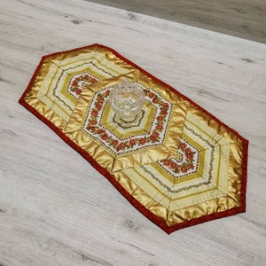 Könnte beinhalten: Dekorativer Tischläufer in Gold und Rot. Der Läufer hat ein Blumenmuster in der Mitte und ist mit goldenem Satin eingefasst. Ein durchsichtiges Glasgefäß befindet sich obenauf.