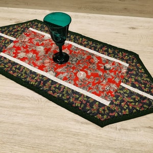 Könnte beinhalten: Ein grüner und roter gesteppter Tischläufer mit weißem Rand und einem Design aus Stechpalmenblättern. Ein grünes Glasgefäß liegt auf dem Tischläufer.