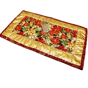 Könnte beinhalten: Ein goldener und roter Tischläufer mit einem Blumenmuster mit Weihnachtssternen. Der Läufer hat einen goldenen Rand und einen gläsernen Kerzenhalter in der Mitte.