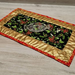 Könnte beinhalten: Ein goldener und roter gesteppter Tischläufer mit einem schwarzen und grünen Stechpalmen-Druck. Der Läufer hat einen goldenen Rand und einen roten und goldenen Besatz. Eine klare Glasschale liegt auf dem Läufer.