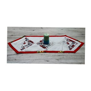 Könnte beinhalten: Ein weißer Tischläufer mit rotem Rand und geometrischem Muster. Der Läufer ist mit roten und grünen Blumendesigns verziert. Eine türkisfarbene Kerze steht auf dem Läufer.