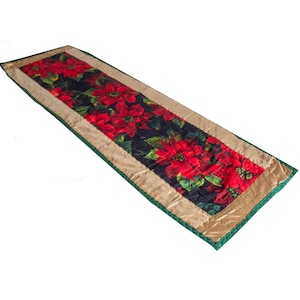 Puede incluir: Un camino de mesa acolchado rojo y verde con estampado de poinsettia. El camino de mesa tiene un borde dorado y un borde verde.