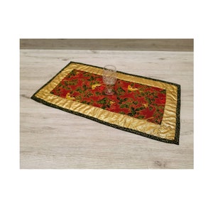 Könnte beinhalten: Ein roter und goldener Tischläufer mit einem festlichen Vogel- und Stechpalmenmuster. Der Läufer hat einen schwarzen Rand und ist gesteppt.