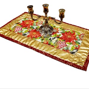 Könnte beinhalten: Ein roter und goldener Tischläufer mit einem weihnachtlichen Blumendruck mit Weihnachtssternen, Vögeln und Ornamenten. Ein goldener Kandelaber mit drei Kerzen steht auf dem Läufer.