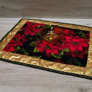 Könnte beinhalten: Ein gesteppter Tischläufer mit schwarzem Hintergrund und einem roten Weihnachtsstern-Blütenmuster. Der Läufer hat einen goldenen Satinrand und einen grünen gewellten Rand.