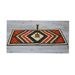 Könnte beinhalten: Ein Tischläufer mit einem geometrischen Muster in Rot-, Braun-, Schwarz- und Goldtönen. Der Läufer ist aus Stoff gefertigt und hat in der Mitte eine Diamantform. Ein goldener Kerzenständer steht auf dem Läufer.