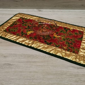Könnte beinhalten: Ein roter und grüner gesteppter Tischläufer mit goldener Einfassung und einer klaren Glasschüssel darauf. Der Läufer zeigt ein festliches Muster aus Stechpalmenblättern und Beeren.