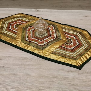 Könnte beinhalten: Ein sechseckiger Tischläufer in Gold und Grün mit einem floralen Muster. Der Läufer ist aus Stoff und hat einen grünen Rand. Ein klares Glasgefäß mit Deckel steht auf dem Läufer.