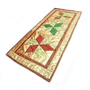 Puede incluir: Un camino de mesa dorado con un patrón de estrellas rojas, verdes y doradas. El camino de mesa está hecho de una tela acolchada con un acabado satinado.