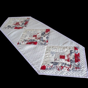 Può includere: Un runner da tavolo bianco con un motivo a rombi con quadrati di tessuto floreale rosso e grigio. Il runner ha un bordo a uncinetto bianco.