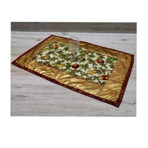 Könnte beinhalten: Ein goldfarbener und roter gesteppter Tischläufer mit einem weihnachtlichen Design mit Stechpalme, Beeren und Ornamenten. Der Läufer liegt auf einer hellbraunen Holzoberfläche mit einem klaren Glas darauf.