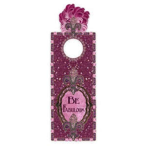 May include: A pink door hanger with a fleur-de-lis design and the text "Be Fabulous". The hanger has a hole for a door knob and is decorated with rhinestones.