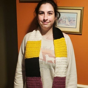 Pode incluir: Uma pessoa vestindo uma camisa branca de manga comprida com uma estampa gráfica em preto e branco. Ela está usando uma echarpe de crochê com listras amarelas, pretas, brancas e roxas.