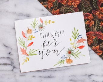 May include: A white card with a watercolor illustration of fall leaves and pumpkins. The card says "Thankful for you" in black calligraphy.