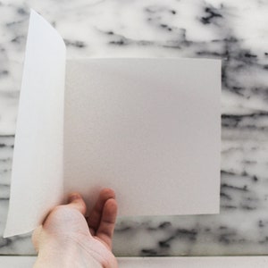 May include: A white sheet of paper is folded in half and held by a hand against a white marble background.