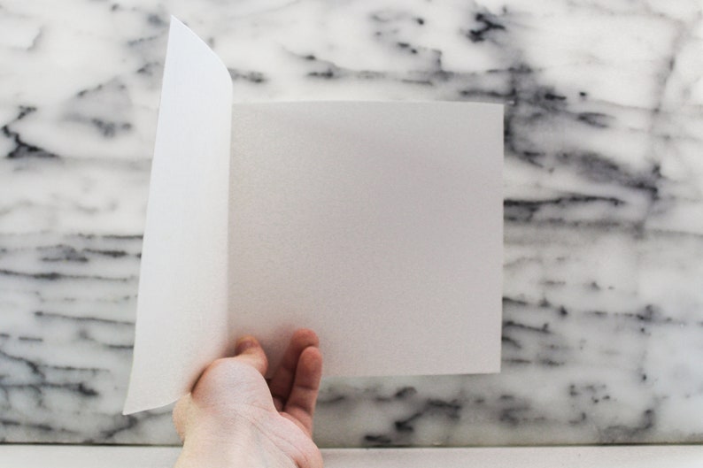 May include: A white sheet of paper is folded in half and held by a hand against a white marble background.