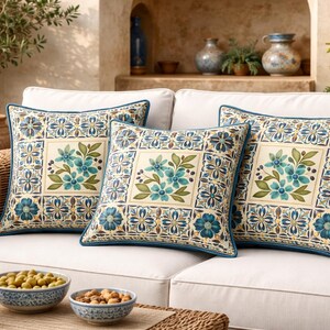 May include: Three square throw pillows with a floral design on a white sofa. The pillows have a cream-colored center with teal flowers and green leaves, surrounded by a blue and cream patterned border. A blue pillow is on the right.