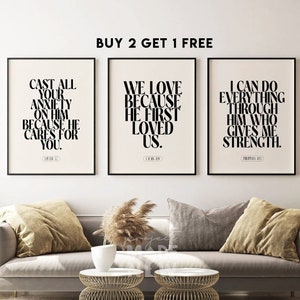 May include: Three black framed prints with white backgrounds and black text. The prints feature inspirational quotes with bible verses. The prints are displayed on a white wall above a gray couch with pillows and a vase of dried pampas grass.