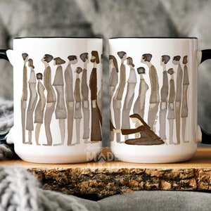 May include: Two white ceramic mugs with black handles. Each mug features a watercolor illustration of a line of people in brown tones. One mug shows a person reaching down to another person in the line. The mugs are on a wooden surface.