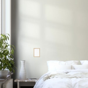 May include: A white bed with white bedding and a white throw blanket. A small wooden frame with a white mat is hanging on the wall above the bed. A vase with a white textured design is on a wooden table next to the bed.