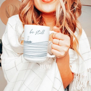 May include: White ceramic mug with black text that reads "be still" and a simple line drawing of a cross and waves. The mug is being held by a person wearing a white sweater.