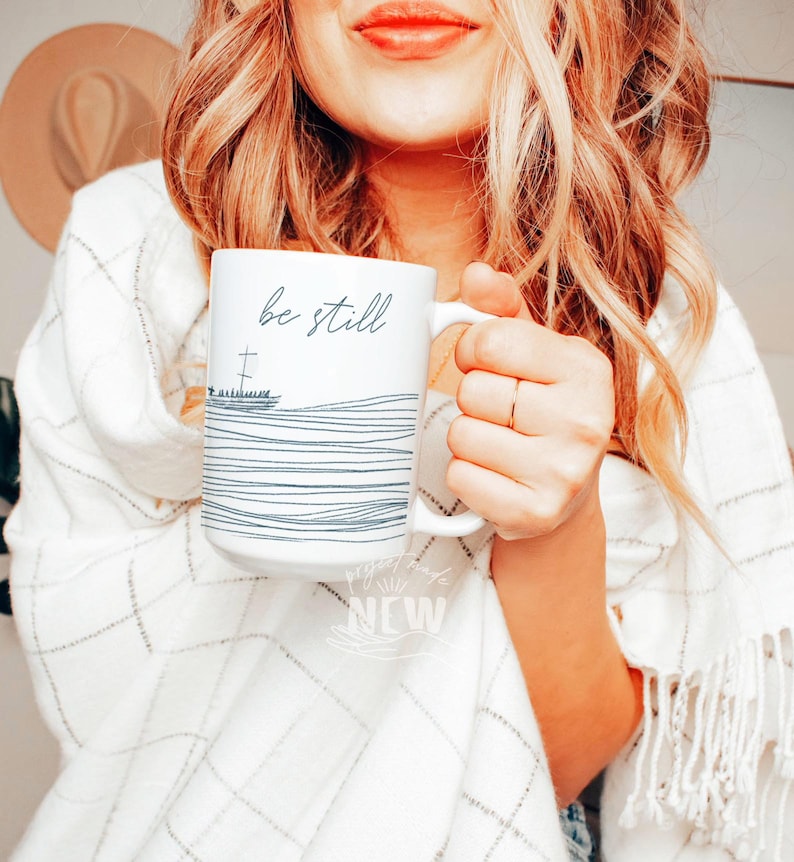 May include: White ceramic mug with black text that reads "be still" and a simple line drawing of a cross and waves. The mug is being held by a person wearing a white sweater.