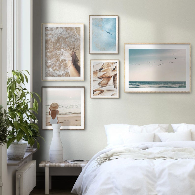 May include: A bedroom with a white bed and a white nightstand. There are five framed pictures on the wall above the bed. The pictures are of a beach scene, a woman in a swimsuit, and a seashell.