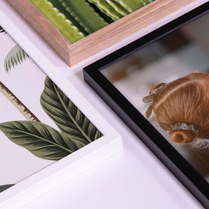 May include: Three different picture frames with different colored frames and images. The frame on the left is white with a tropical leaf pattern. The frame in the center is light brown wood with a green cactus pattern. The frame on the right is black with a photo of a girl's back.