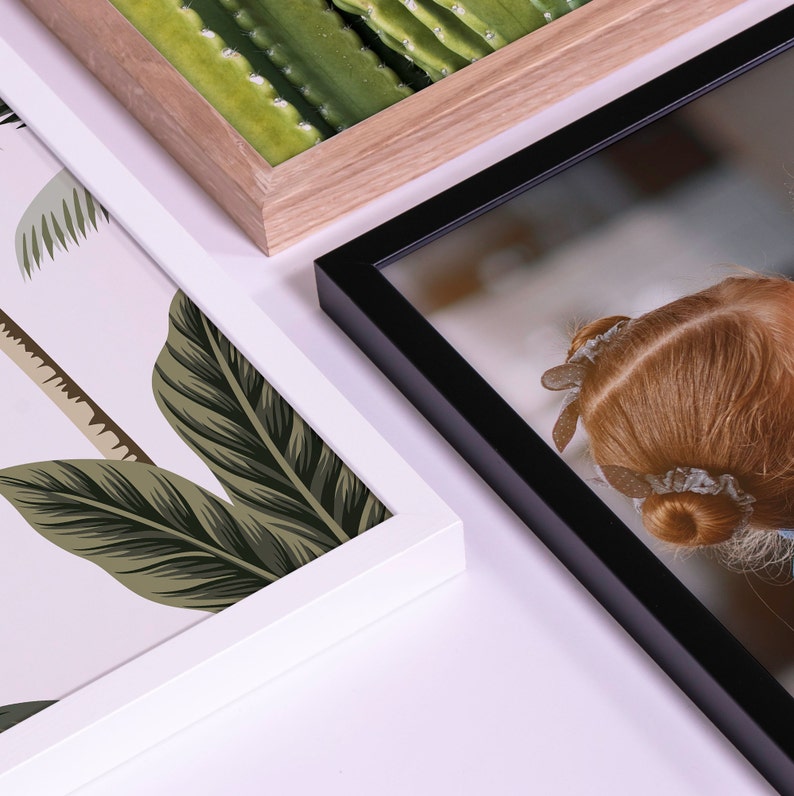 May include: Three different picture frames with different colored frames and images. The frame on the left is white with a tropical leaf pattern. The frame in the center is light brown wood with a green cactus pattern. The frame on the right is black with a photo of a girl's back.