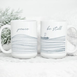 May include: Two white ceramic mugs with black text and a wavy line design. The mug on the left says "peace" and the mug on the right says "be still".
