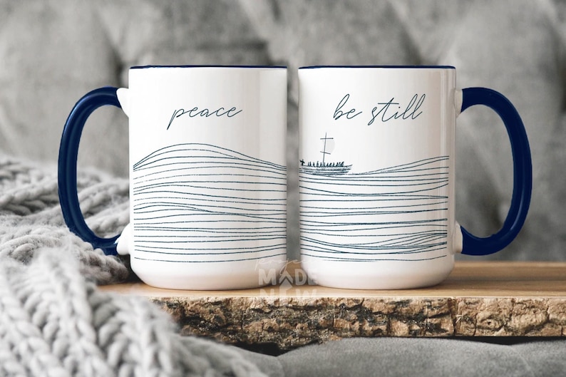 May include: Two white ceramic mugs with blue rims and handles. The mug on the left says "peace" and the mug on the right says "be still". Both mugs feature a blue and white wavy line design.