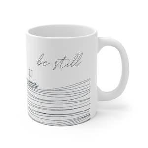 May include: White ceramic mug with a black and white illustration of a boat on a wavy line. The text "be still" is written in black cursive on the mug.