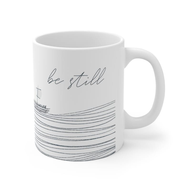 May include: White ceramic mug with a black and white illustration of a boat on a wavy line. The text "be still" is written in black cursive on the mug.